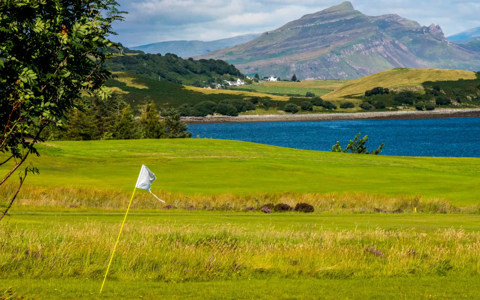 Scottish golf course with white flag on green fairway, surrounded by lake, hills, and mountains under cloudy sky.