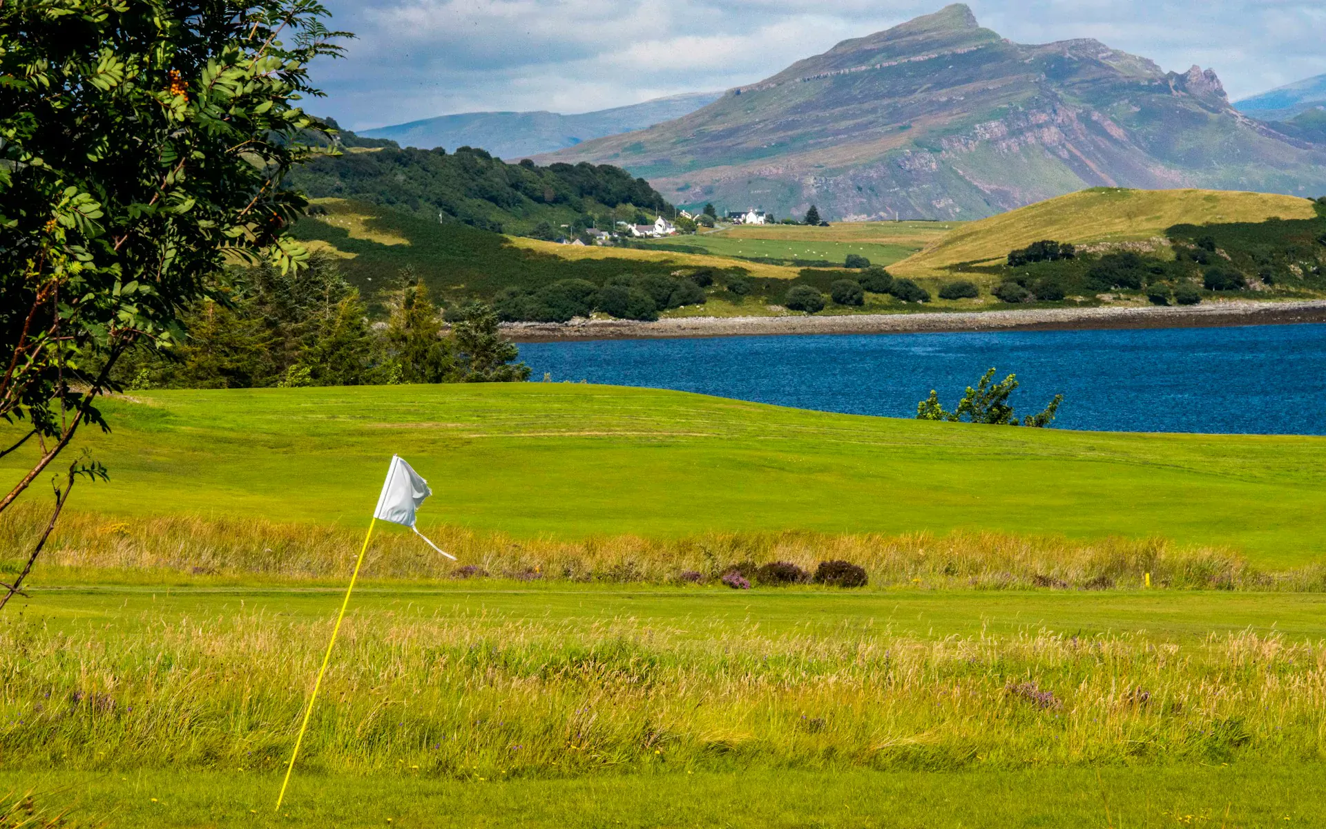 Scottish golf course with white flag on green fairway, surrounded by lake, hills, and mountains under cloudy sky.
