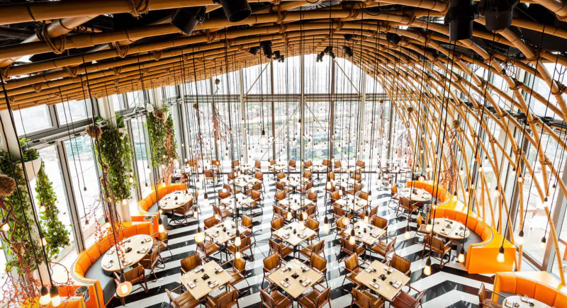 Aerial view of SushiSamba restaurant with bamboo ceiling, orange seating, round tables, and city skyline through glass walls.