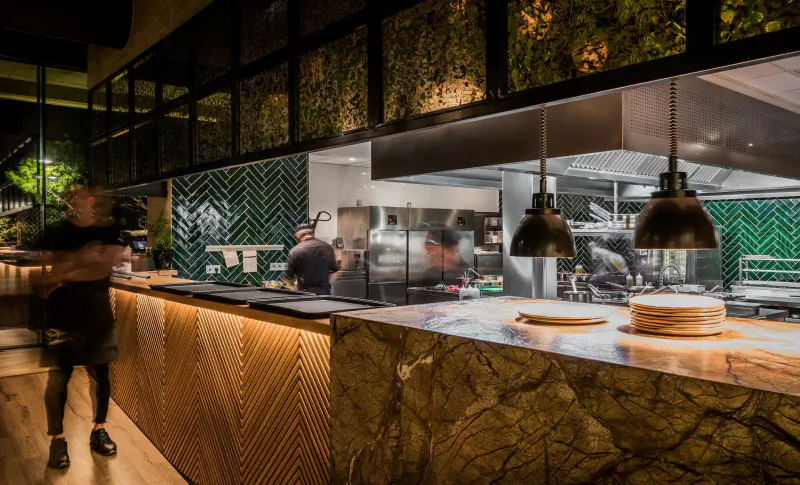 Blurry chefs working in modern Breathe Marbella kitchen with green tiles, pendant lights, and stacked plates on wooden counter.