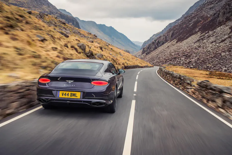 Rear view of black Bentley GT Mulliner Blackline speeding on winding mountain road, UK plate VRM 644, rocky valley backdrop.