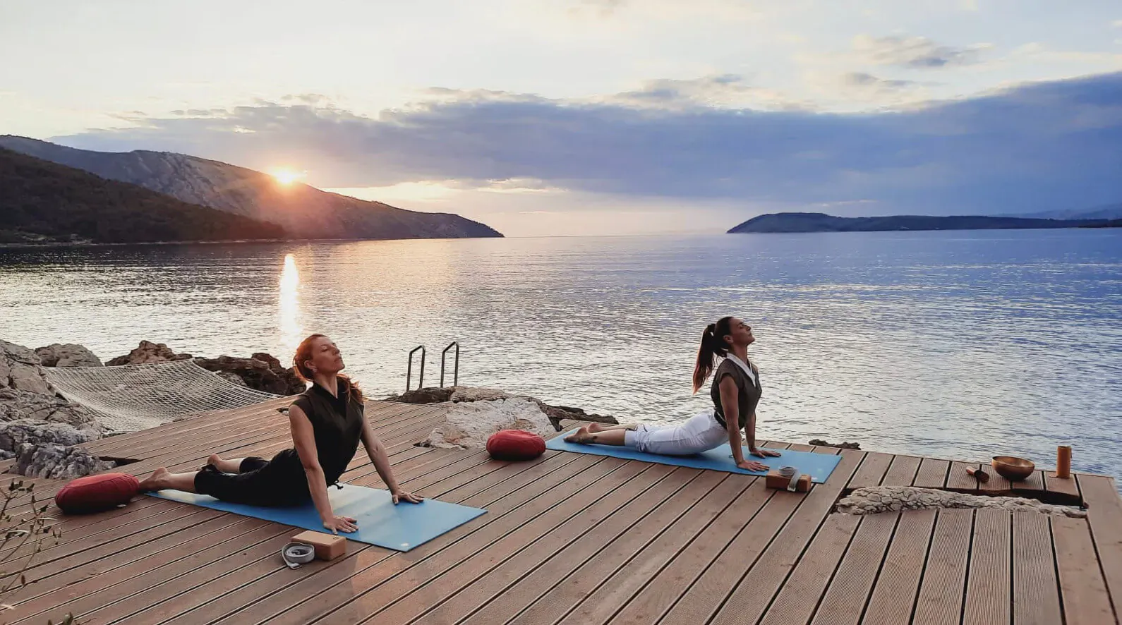 Two women practicing yoga on blue mats on a wooden dock at sunset, overlooking sea and mountains at Maslina Resort spa.