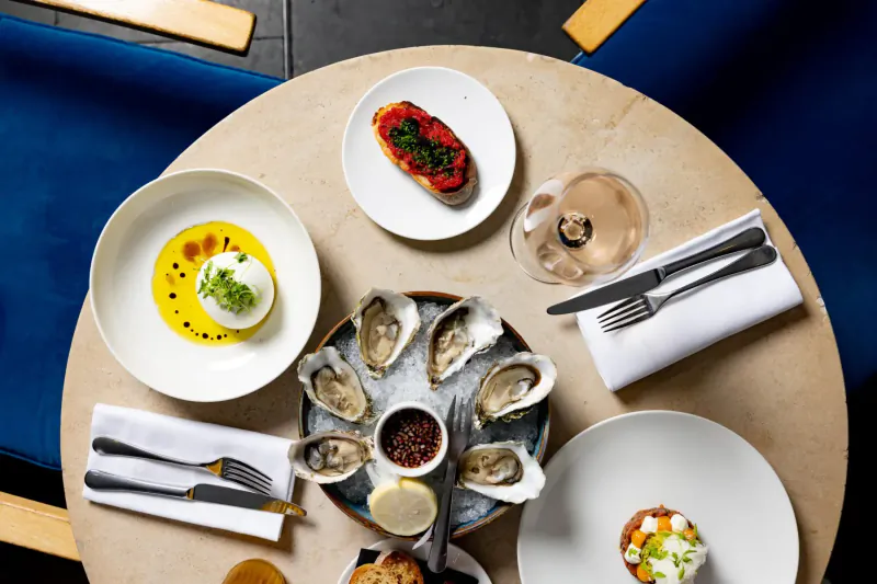 Elegant table at Bluebird Chelsea with oysters, burrata, tomato tart, and rosé glass for festive season.