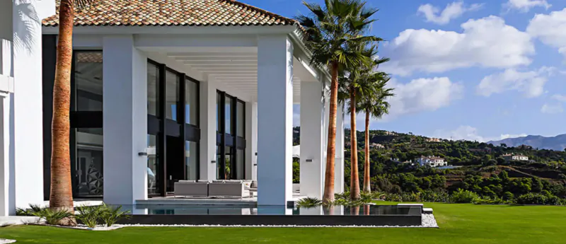 Modern white luxury villa with large glass windows, palm trees, infinity pool, and Marbella mountain views.
