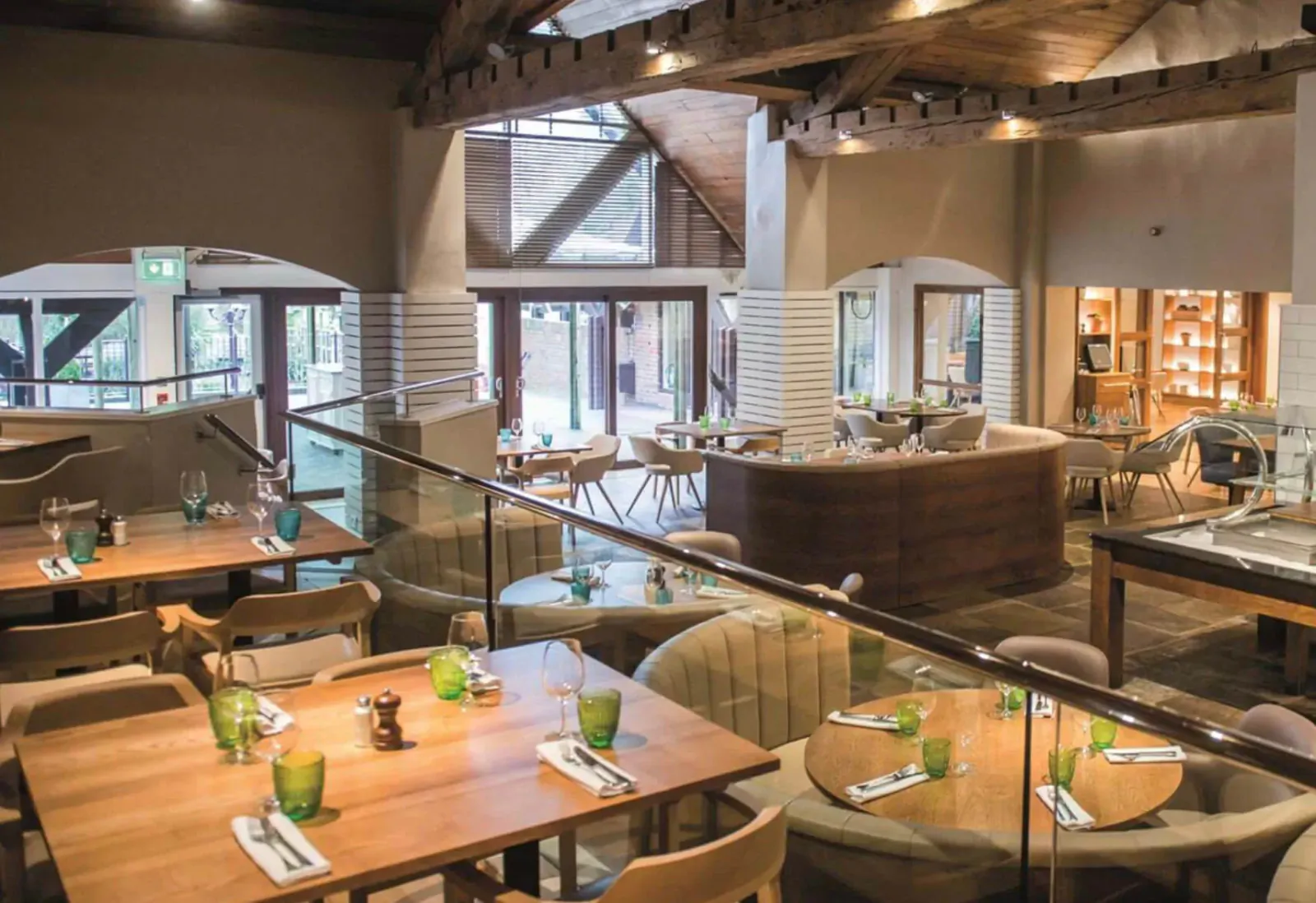 Spacious modern hotel restaurant with wooden beams, glass tables with green glassware, and large windows overlooking greenery.