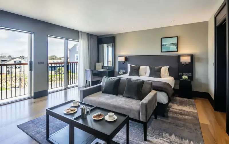 Spacious modern hotel suite with king bed, gray velvet sofa, coffee service on table, balcony view of gardens.