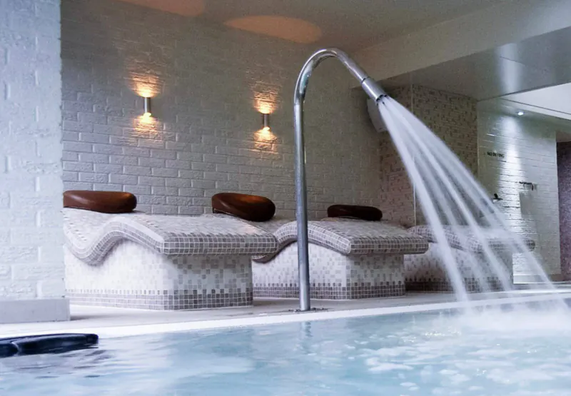 Luxury spa with curved shower spraying water over two mosaic lounge chairs by indoor pool at Moddershall Oaks.