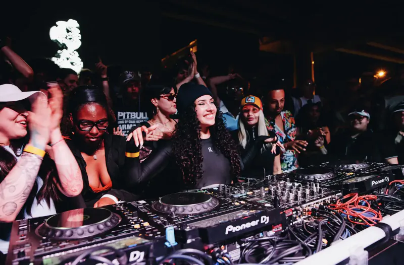 DJ with curly hair in black bodysuit performs energetically on Pioneer DJ decks at crowded Boiler Room Miami club event.