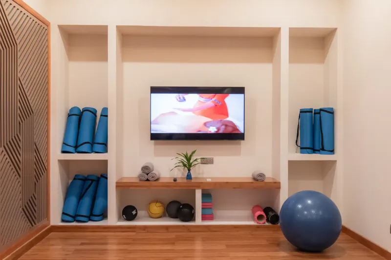 Modern resort gym with blue yoga mats, exercise balls, weights on shelves, and wall-mounted TV.