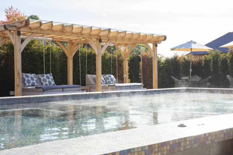 Wooden pergola with blue cushioned swings over steaming hot tub at spa, yellow umbrellas and hedges in background.