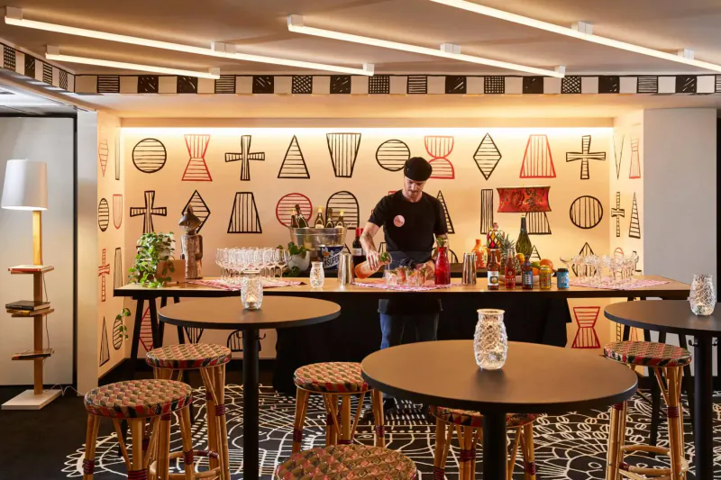 Bartender mixing drinks at modern bar with geometric wall art, Halloween gallery vibe, round tables and stools