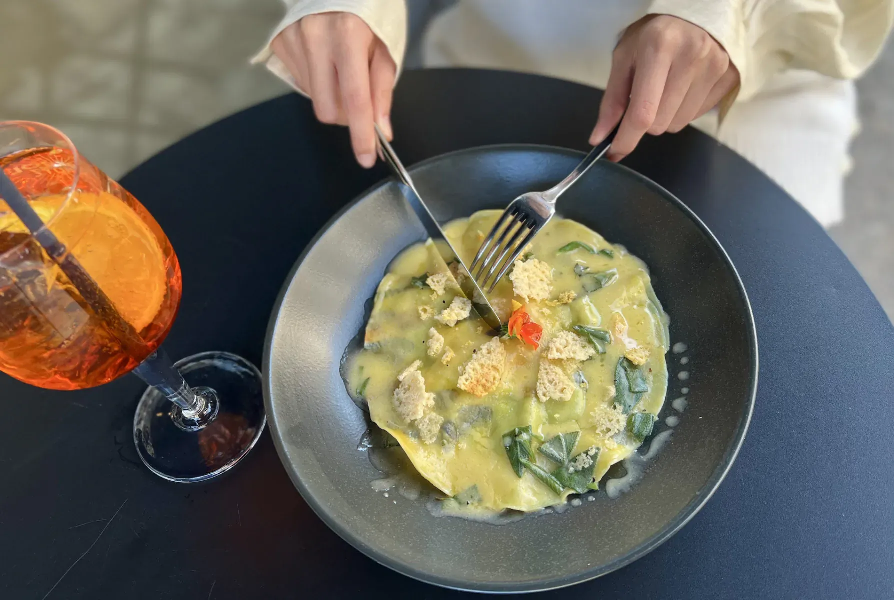 Hands twirling fork in bowl of yellow creamy spinach ravioli with orange Aperol Spritz on table