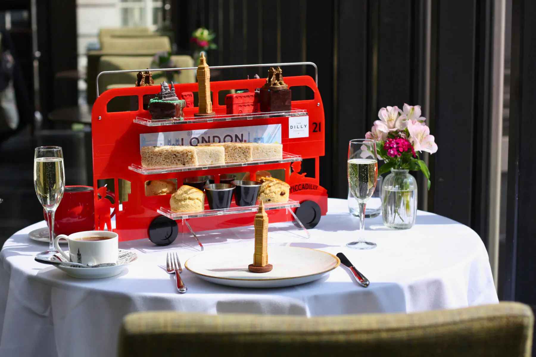 Red double-decker bus-shaped tiered stand with sandwiches, scones, cakes, champagne flutes, and tea on white tablecloth for London afternoon tea.