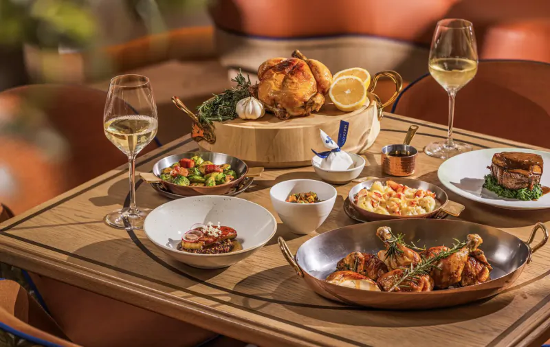 Elegant dining table with roasted chicken, salads, seafood, meats on wooden board, garnished with lemon, in upscale setting.