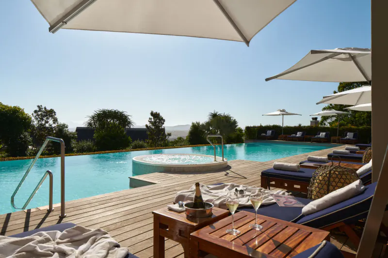 Luxury infinity pool deck at Delaire Graff Estate with champagne, wine glasses, loungers, and Cape Winelands views under white umbrellas.