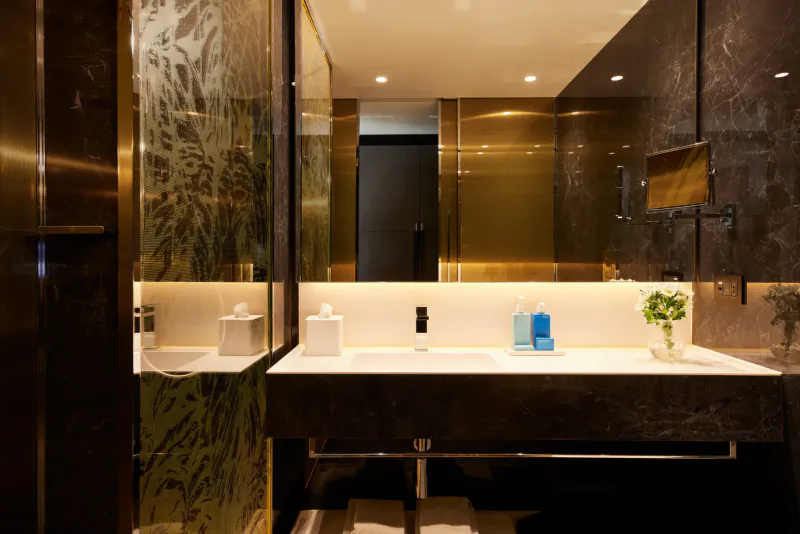 Luxurious modern hotel bathroom with black marble vanity, gold fixtures, blue soap dispenser, plants, and mirrored walls.