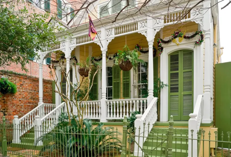 Ornate yellow Victorian house with green shutters, purple flag, garlands, plants, and white steps in lush garden.