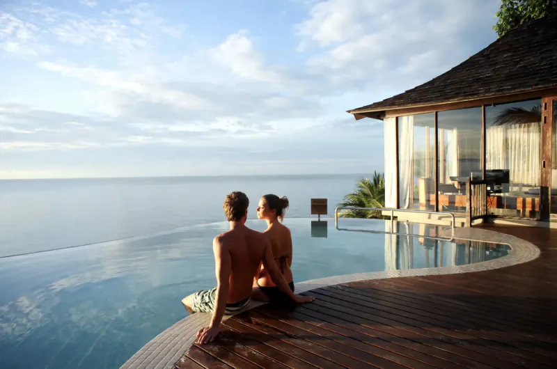 Couple sits embracing on edge of infinity pool at sunset, overlooking ocean from luxurious wooden deck resort.