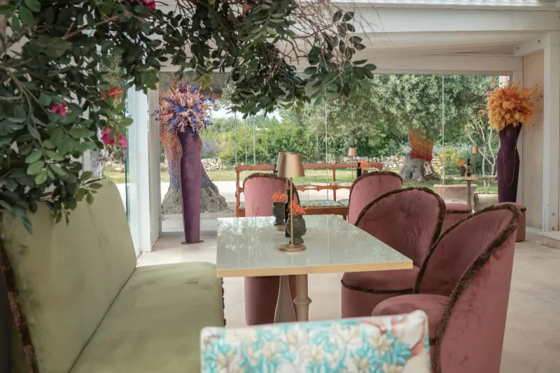 Elegant outdoor seating at Zahir Country House with green sofa, pink velvet chairs, glass table, and vibrant floral arrangements under trees.