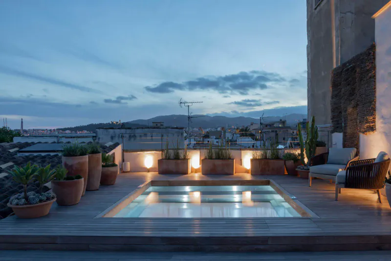 Twilight rooftop pool at Can Bordoy Grand House & Garden, lit with underwater lights, surrounded by terracotta pots, agaves, chairs, and distant mountains.