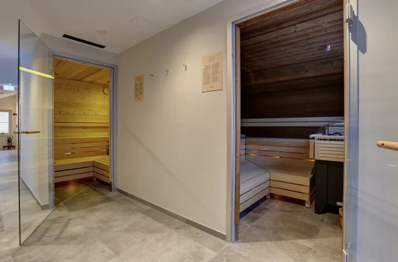 Two adjacent wooden saunas with benches in a modern hotel spa hallway, glass doors and tiled floor.