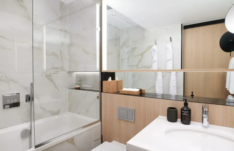 Modern hotel bathroom with glass shower, bathtub, white vanity, wood accents, and towels in Nobu Hotel Marylebone.