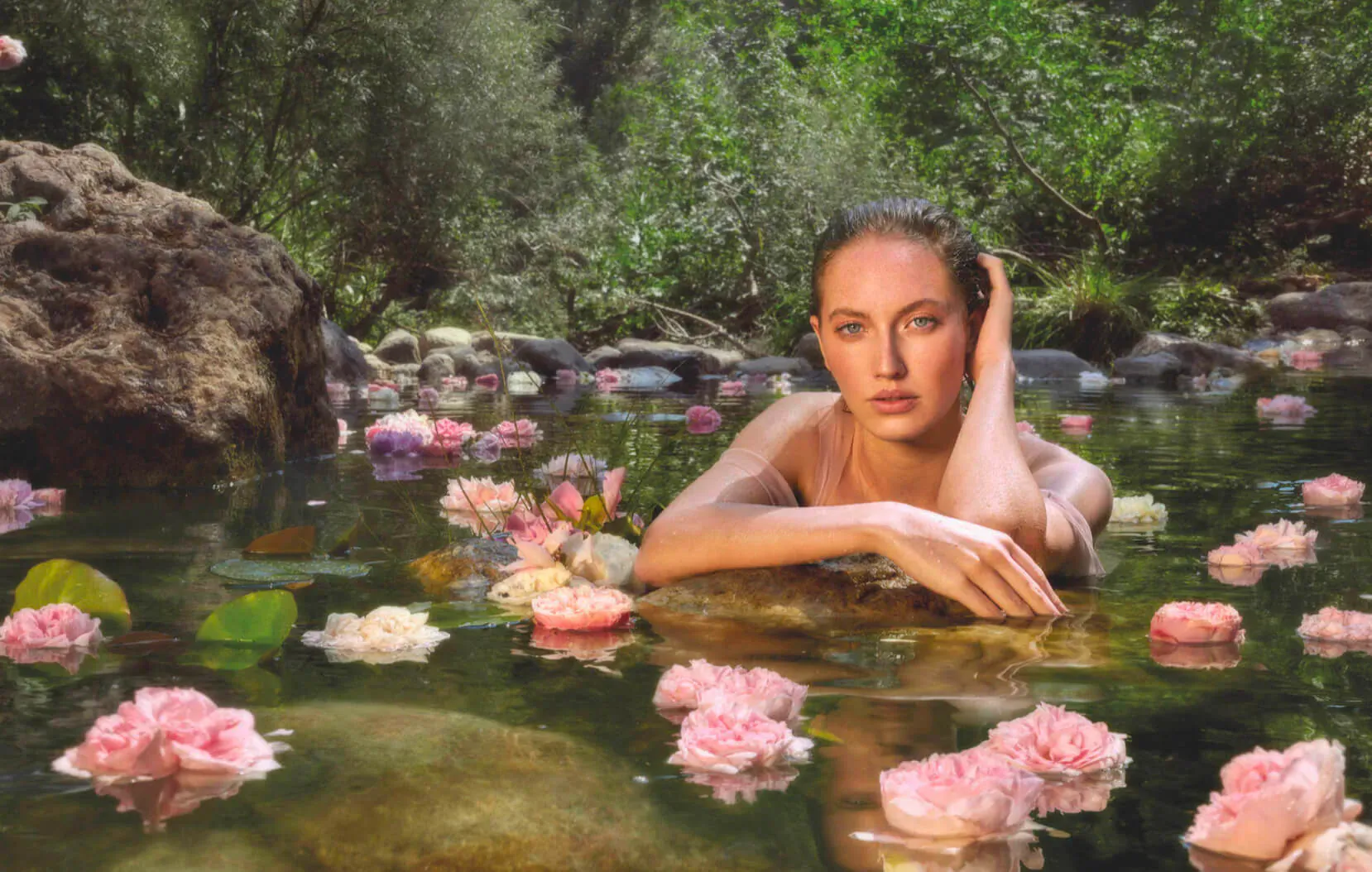 Nude woman with hand on cheek, reclining in lush green forest stream surrounded by pink rose petals