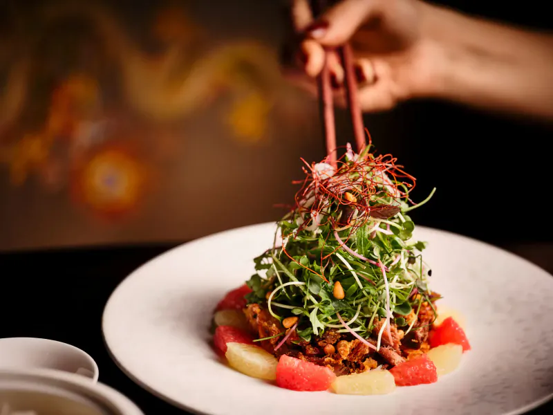 Hand with chopsticks lifting sprouts from a modern Cantonese salad of greens, watermelon, and apple on white plate