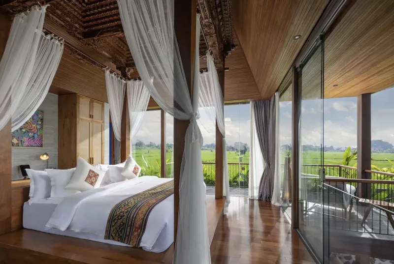Luxurious Balinese bedroom with canopy bed, white drapes, and panoramic rice field views through glass doors.