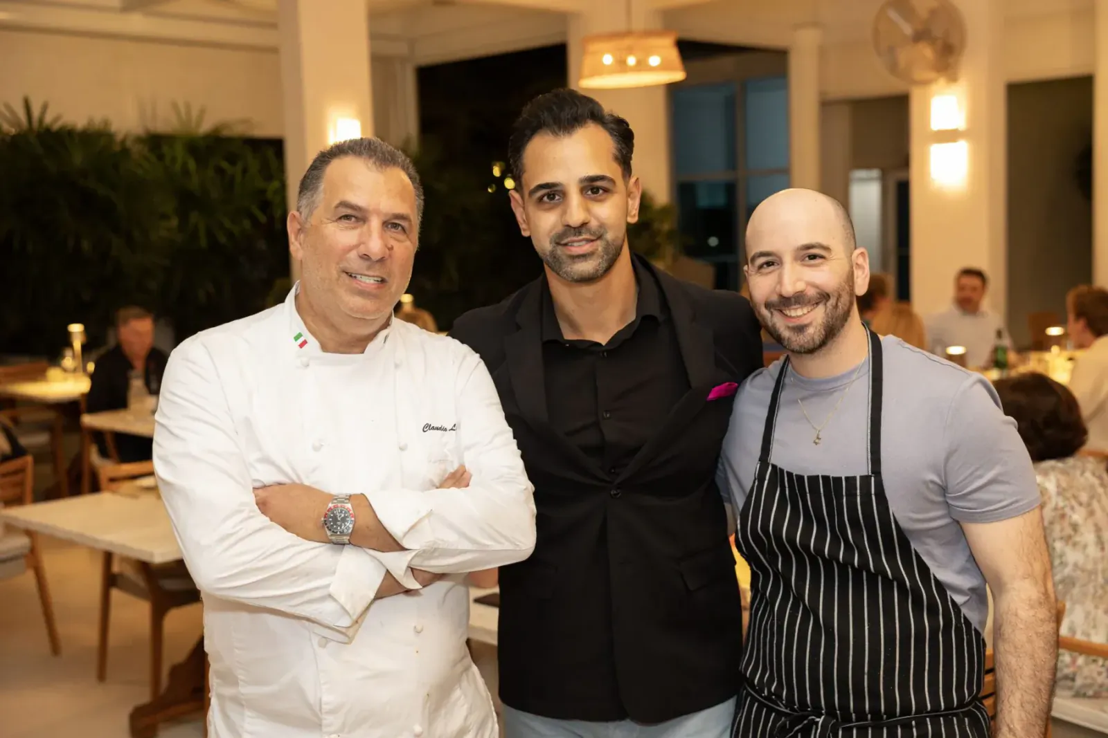 Three smiling men posing together in a restaurant: chef in white uniform, man in black shirt, man in gray tee and apron.