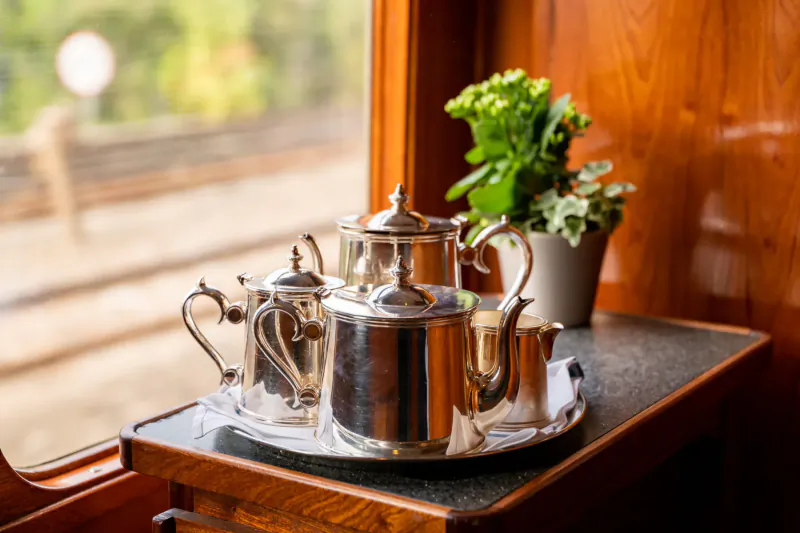 Silver vintage tea set with teapot, cream jug, sugar bowl on tray, green plant, luxury train wooden interior window view.