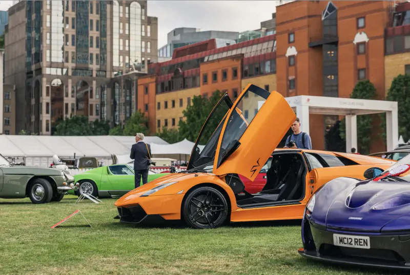 Orange Lamborghini with doors up at London Concours 2024, beside purple McLaren and green MG on grass amid city buildings.