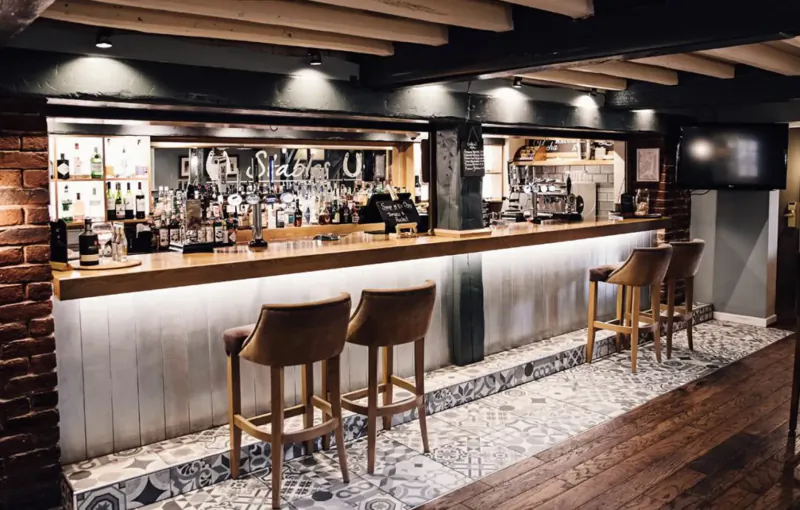 Elegant wooden bar at Manor House Hotel with backlit shelves of bottles, high stools, brick walls, and tiled floor.