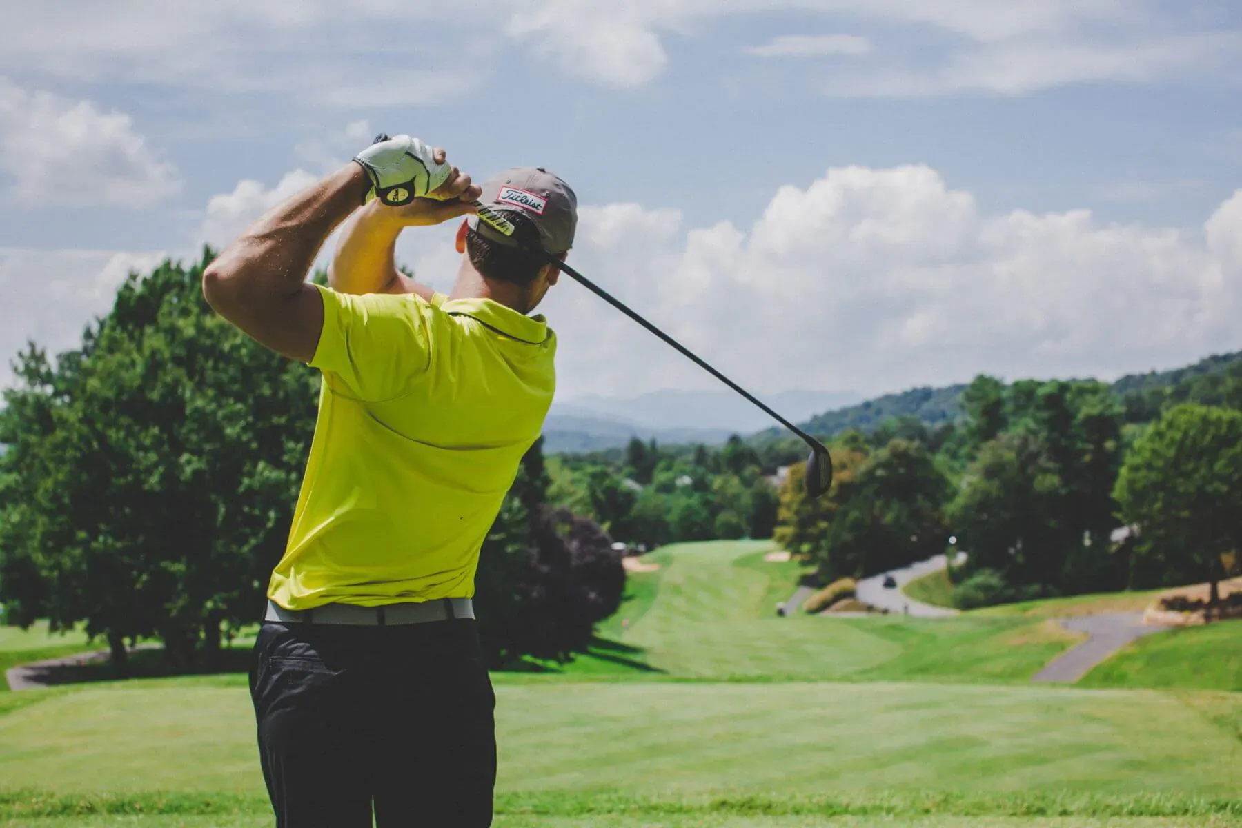 Man in yellow shirt swings golf club on lush green course with trees and mountains under cloudy sky
