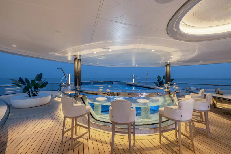 Luxury yacht 'Kismet' outdoor bar with white stools around glowing counter, pool, plants, ocean view at dusk