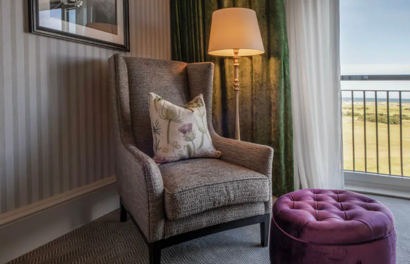Elegant gray wingback armchair with floral pillow and purple ottoman beside lamp in luxury hotel room with sea view.