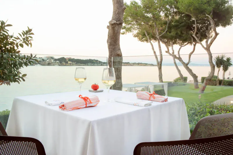 Elegant outdoor table set with white linens, orange napkins, white wine glasses, and cherry tomato, overlooking lake at sunset amid pine trees.