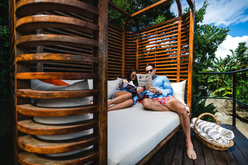 Couple relaxing on bed in wooden gazebo at 21 Nettleton Boutique Hotel, man reading newspaper, Cape Town mountain-sea view.