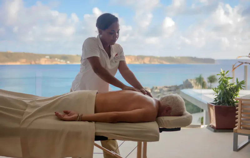 Female therapist giving massage to shirtless older man on table, ocean view balcony at luxury resort.