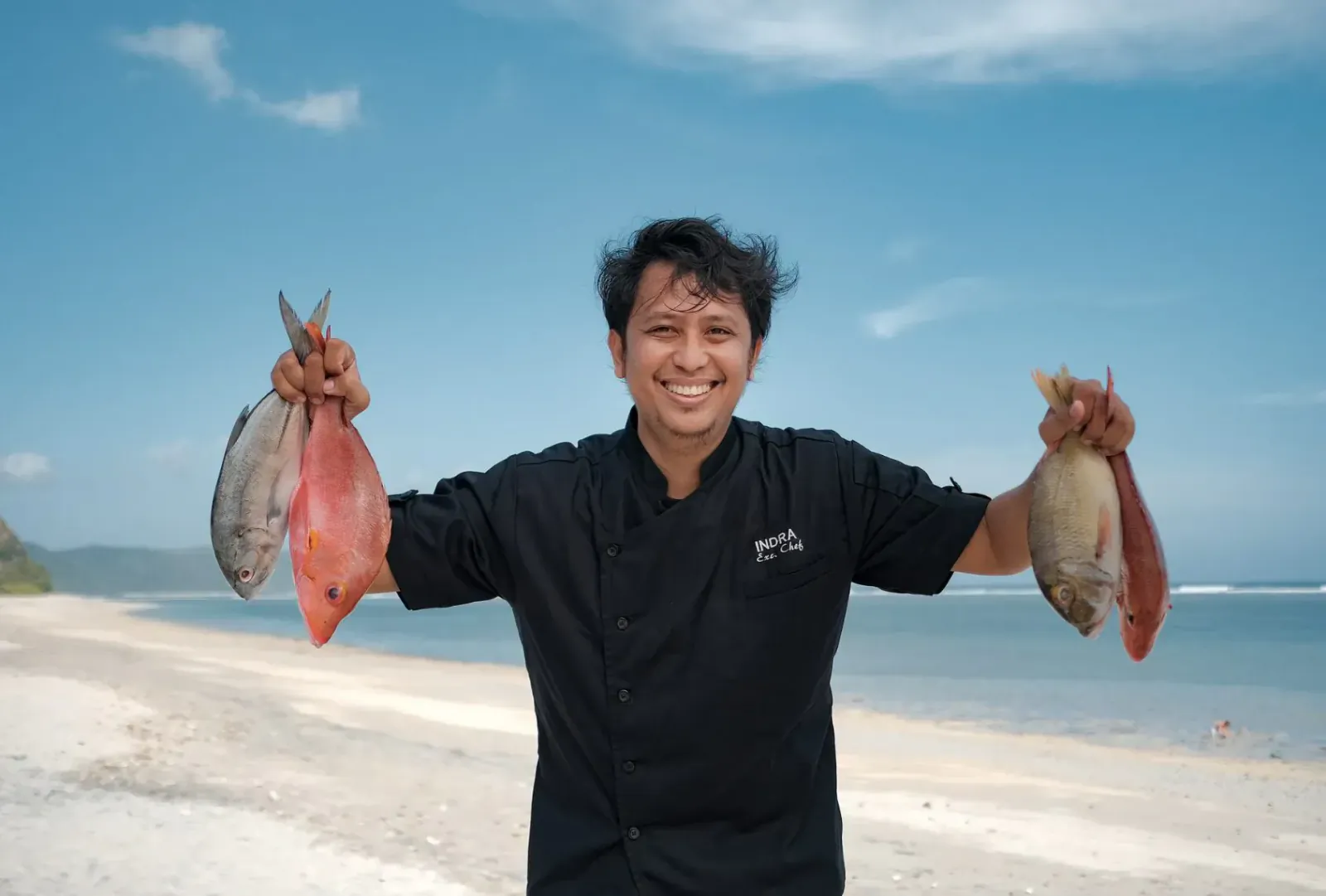 Smiling chef in black uniform holding up red snapper fish on Amber Lombok beach with ocean view
