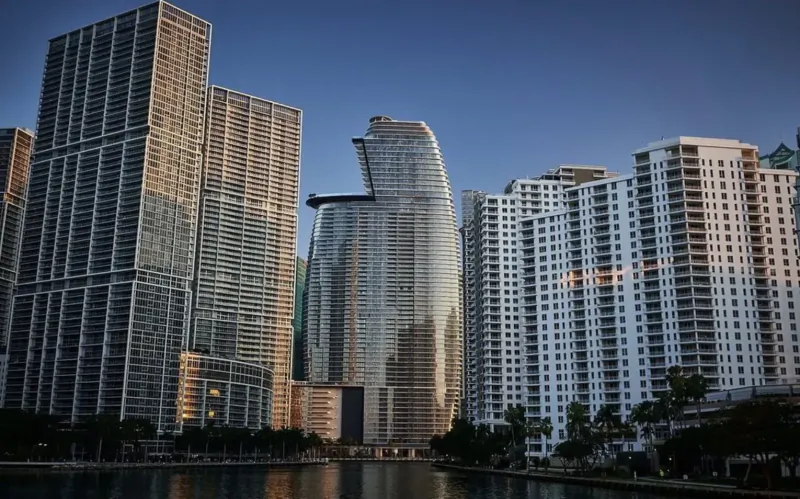 Aston Martin Residences Miami: curved glass skyscraper amid modern high-rises at sunset by waterfront.