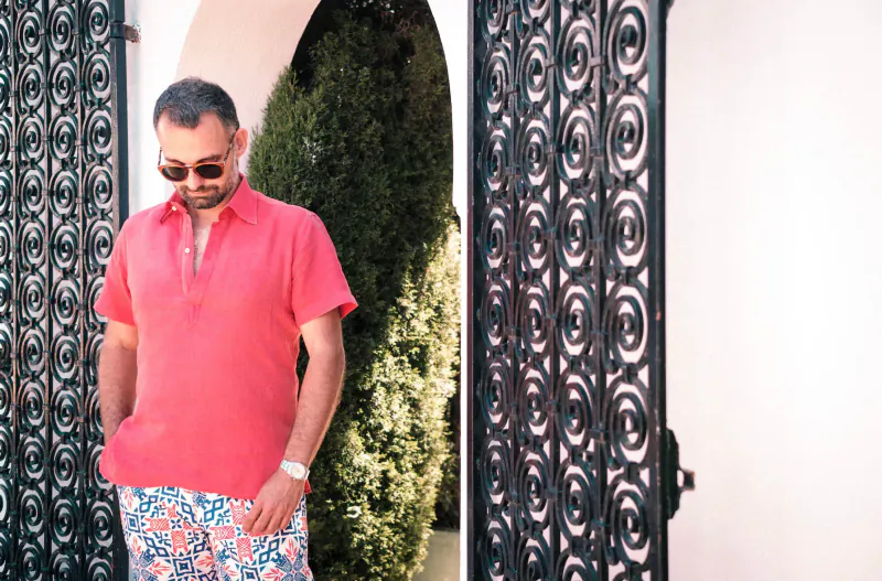 Man in pink polo and floral shorts stands by ornate black gate in Marbella archway, sunglasses on.
