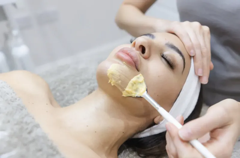 Esthetician applying yellow facial mask with spatula to client's face during spa treatment