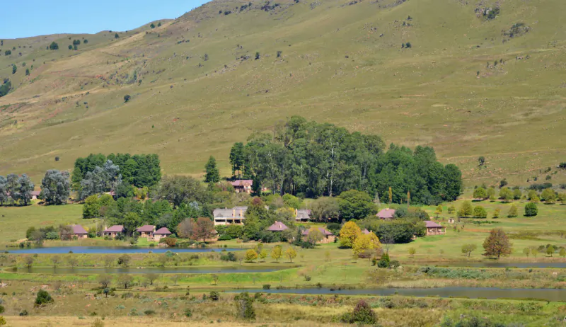 Aerial view of a scenic rural retreat with cluster of buildings, trees, river, and rolling green hills under blue sky.