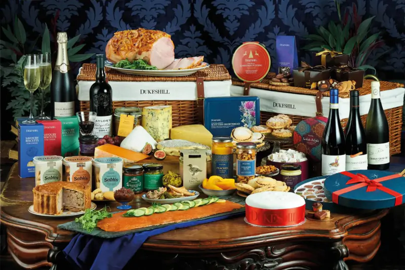 Festive Christmas hamper on ornate table: roast ham, salmon, cheeses, biscuits, wine, champagne, 'Dukeshill' baskets.