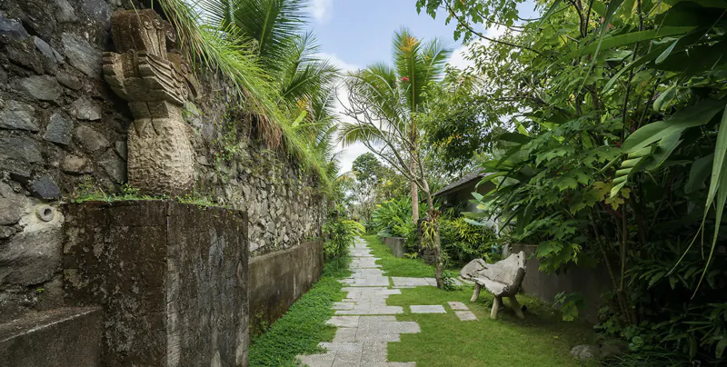 Stone winged statue on mossy wall beside lush tropical garden path and bench, Bali style