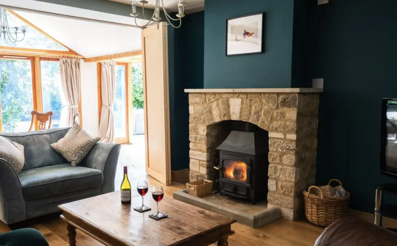 Cozy Cotswolds living room with stone fireplace, lit fire, blue sofa, coffee table with red wine glasses and bottle, garden view.
