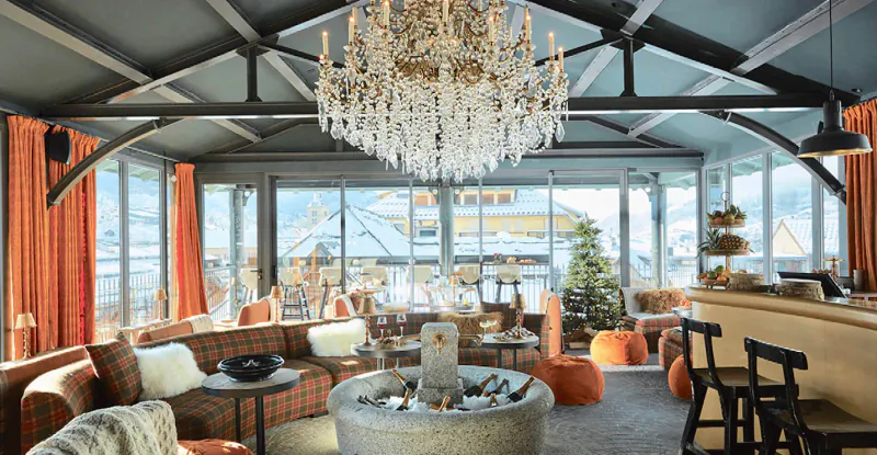 Elegant Megève lounge with chandelier, orange seating, bar, and snowy mountain views through glass walls.