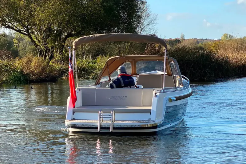 Rear view of white luxury motorboat with beige bimini top and UK flags cruising on calm river amid autumn trees.