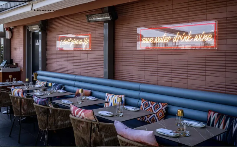 Outdoor rooftop bar with neon signs 'Come water drink wine', blue banquette seating, colorful cushions, and set tables.